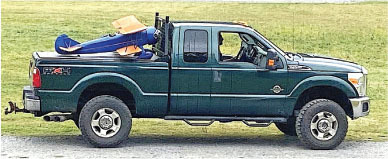 Green pickup truck carrying a large toy plane in the bed, parked on gravel.