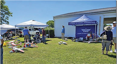 People gather near model planes under tents on a sunny lawn.