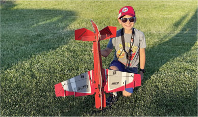 Boy kneels on grass, smiling with a red and gray toy airplane, wearing a red cap and sunglasses.