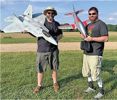 Two men smiling, holding model fighter jets in a grassy field.