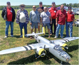 Eight men standing behind a large model airplane on grass.
