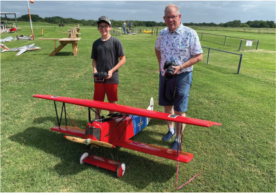 Teen and older man with red model biplane on grassy field.