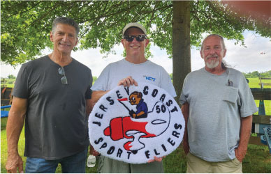 Three men outdoors, one holding a Jersey Coast Sport Fliers logo sign.