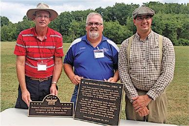 Three men stand outdoors holding commemorative plaques.