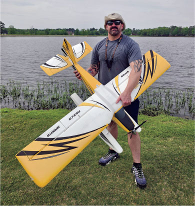 Man holding a large yellow model airplane by a lake.