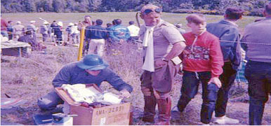 Group of people outdoors, some in costumes, with a man rummaging in a box labeled "AXL-2650".