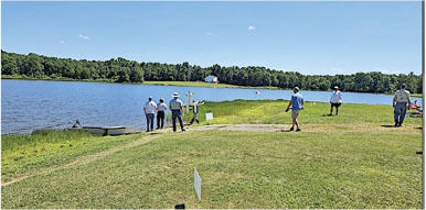 People walking near a lakeside on a sunny day with trees in the background.