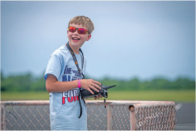 Boy with red sunglasses smiling, holding a drone controller outdoors.