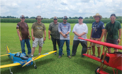 Seven men standing on grass with model airplanes, blue and red, under a cloudy sky.