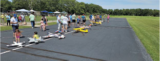 People gathered on a paved area with model airplanes on a sunny day.