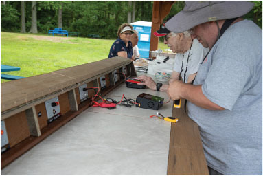 People working at an outdoor electronics bench, surrounded by trees and grass.