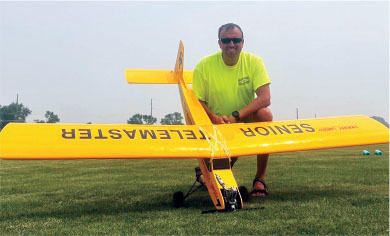 Man kneeling behind a large yellow model plane on grass, wearing sunglasses and a bright shirt.