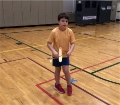 Child in gym holding a paper plane, wearing orange shirt and blue shorts.