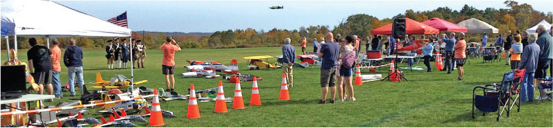 A field event with model airplanes, people watching, tents, and orange cones.