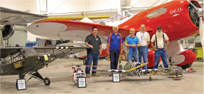 Five men stand in front of model planes and a large red aircraft.