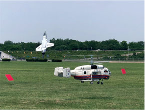 Helicopter flying over grass near parked jet and red markers.
