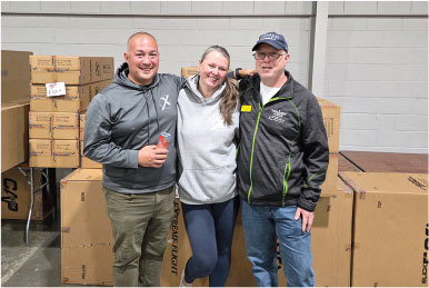 Three people smiling, standing in front of stacked cardboard boxes.