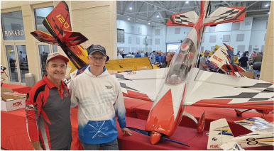 Two people posing with a large red and white model airplane indoors.
