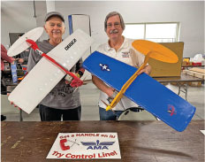 Two smiling people holding model planes indoors, with AMA flyers on the table.