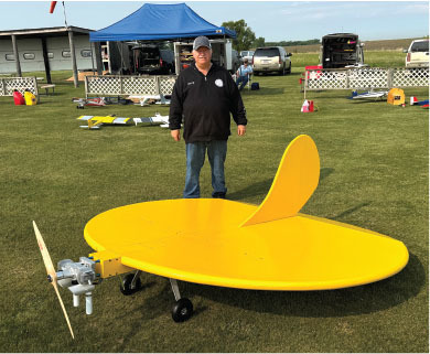 Man standing behind a large yellow model airplane on a grassy field.