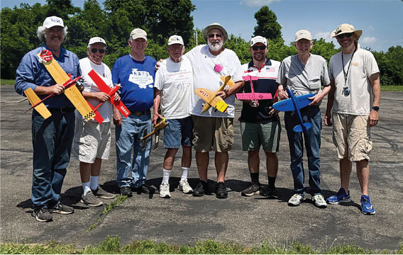 Eight men holding colorful model planes, standing outdoors.