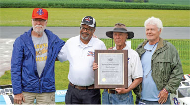 Four men outdoors, one holding a framed certificate, in casual attire.