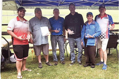 Seven men standing under a canopy, holding awards, on a grassy field.