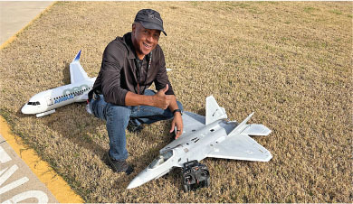 Man kneeling on grass with two model planes, smiling, giving thumbs up.