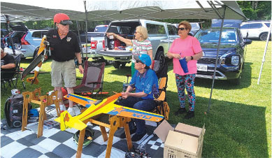 Four people with model planes under a canopy in a grassy field.