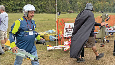 Man with helmet and model plane walks beside person in Darth Vader costume outdoors.