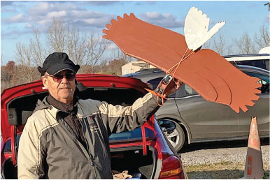 Man holding a large model bird beside a parked car.