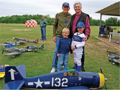 Four people smiling by model airplanes on grass. Child in baseball uniform.