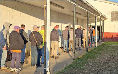 People standing in line outside a building in sunlight.