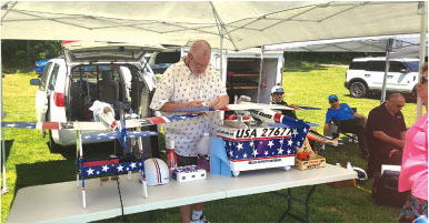 Man working on model plane, USA-themed, under canopy; others seated nearby.