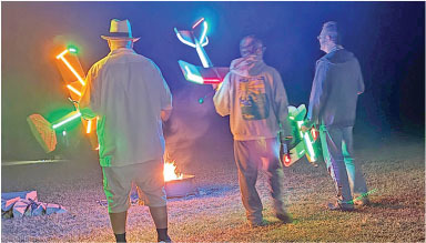 Three people holding illuminated toy planes near a campfire at night.