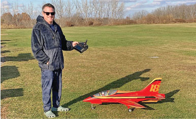 Man with a remote controls a red model jet on grass.
