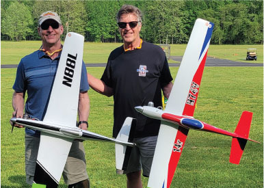 Two men smiling, holding model planes on a grassy field.