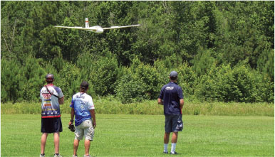 Three people facing a flying model plane in a green field.