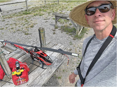 Man with straw hat and sunglasses poses with red model helicopter on wooden table outdoors.