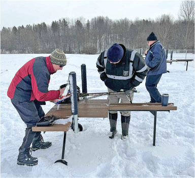 Three people in winter clothing assemble equipment on a snowy field.
