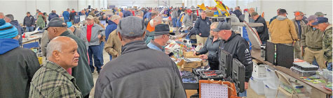 Crowded indoor market with people browsing tables filled with items.