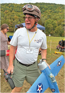 Man holding blue model airplane, wearing helmet and goggles, on grassy field.