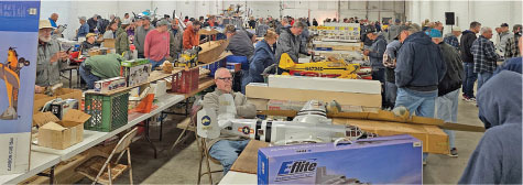Crowded indoor event with model airplanes on display tables.