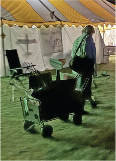 Man pushing a cart under a tent with chairs and bags.