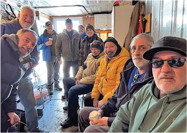Group of men smiling indoors, some seated, wearing winter jackets and hats.