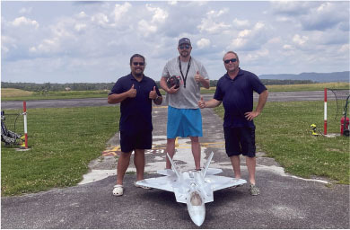 Three men pose on a runway with a model jet, giving thumbs up.