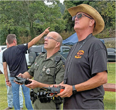 Two men flying drones outdoors, one in uniform, one in a straw hat.