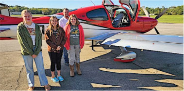 Four people stand smiling in front of a red and white small aircraft on a sunny day.
