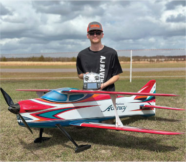 Person holding a remote, standing behind a large model airplane on grass.