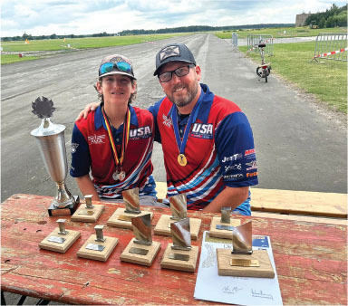 Two individuals in red USA shirts with medals, sitting behind numerous trophies on a table.